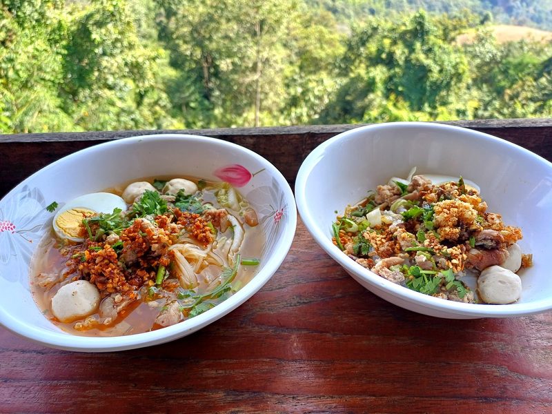 ก๋วยเตี๋ยวต้มยำสองชามวางบนราวระเบียง มีป่าเขียวเป็นฉากหลัง