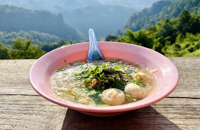 ก๋วยเตี๋ยวน้ำใสใส่ลูกชิ้นในชามสีชมพู วางบนเคาน์เตอร์ไม้หน้าวิวภูเขา