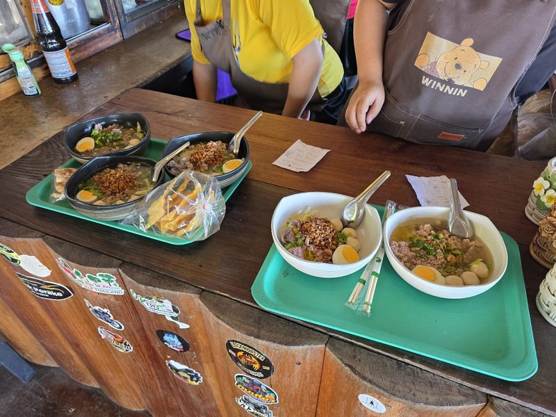 ชามก๋วยเตี๋ยววางบนถาดสีเขียวที่เคาน์เตอร์สั่งอาหารของร้าน